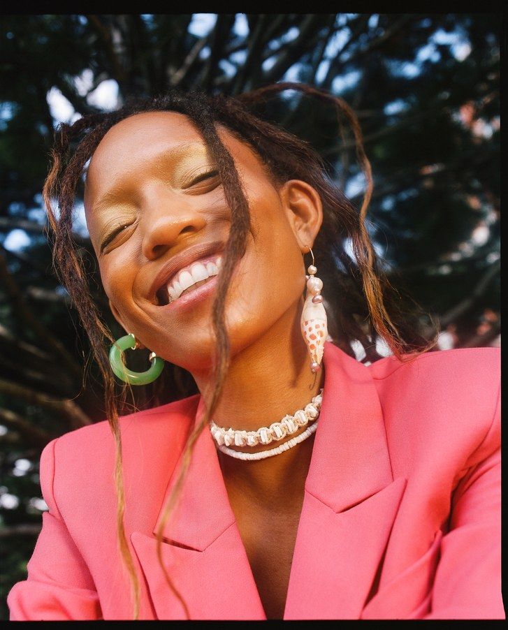 Adesuwa Aighewi pictured smiling in a pink blazer, one jade earring, a shell earring, and a matching choker
