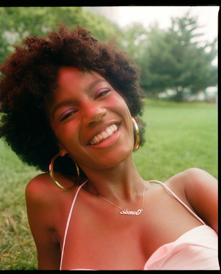 Ebonee Davis pictured laying down on the grass, smiling with a pink top and large gold hoops with a curly fro. 