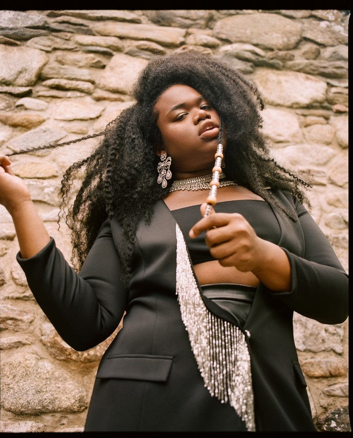 jari Jones poses with beaded strand in hand. They also are wearing a black blazer with hanging beaded detail, matching embellished earrings and necklace in front of a stone wall