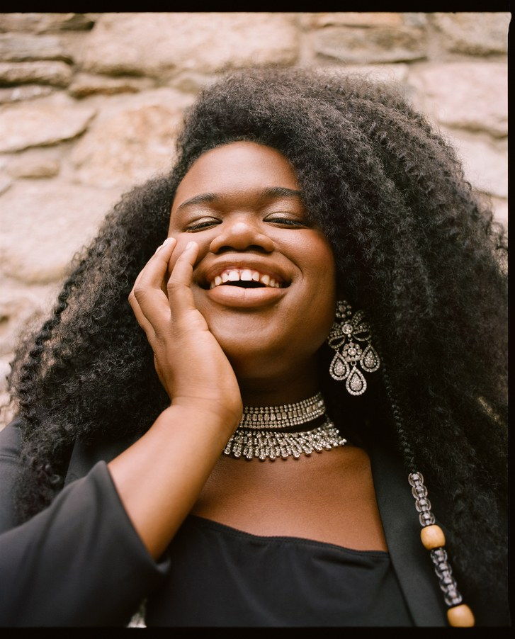 jari Jones pictured laughing with their hand on their face in a black blouse and matching blazer. They wear long, thick, kinky hair with a beaded strand that sticks out. 