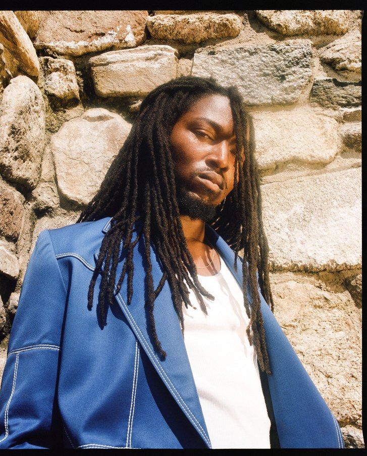 Mikelle Street pictured in front of a stone wall wearing a cobolt blue blazer, a white tank top, thin gold chains, and locs